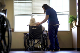 A caregiver gently places a hand on a wheelchair-bound older persons shoulder near a sunlit window.