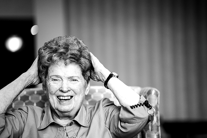 Elderly woman with short hair laughing, hands on head, sitting on a patterned chair.