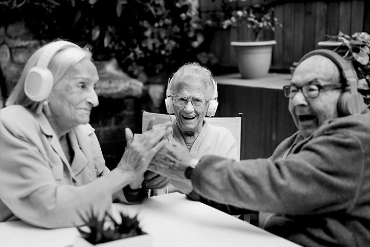 Three elderly people wearing headphones are joyfully clapping hands together at an outdoor table.