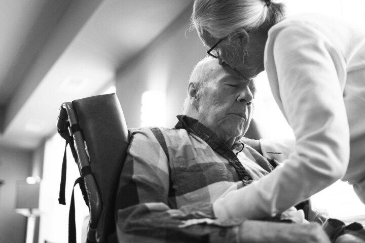 In a brightly lit room, an elderly man in hospice care receives a tender forehead kiss from a woman.