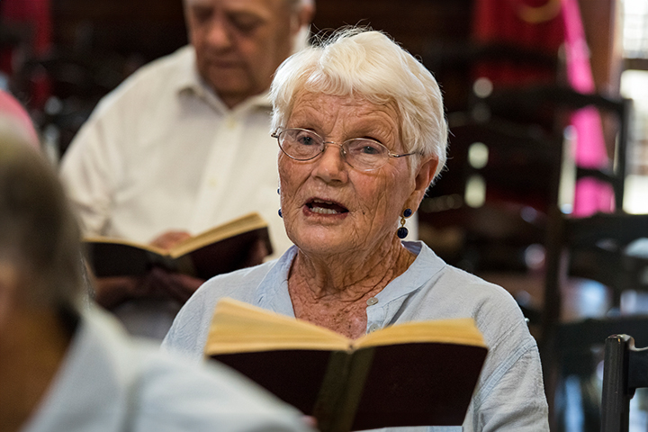 Elderly woman with white hair and glasses singing from a book during a spiritual program, seated indoors.