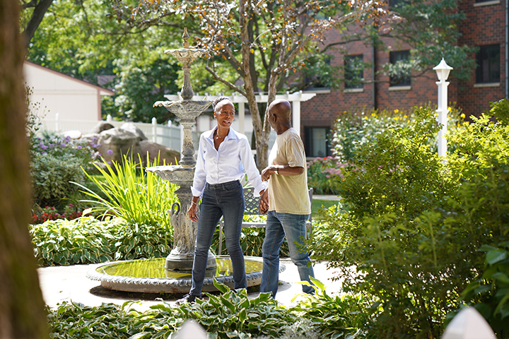 Two people walk together by a decorative fountain in a lush, green garden on a sunny day.