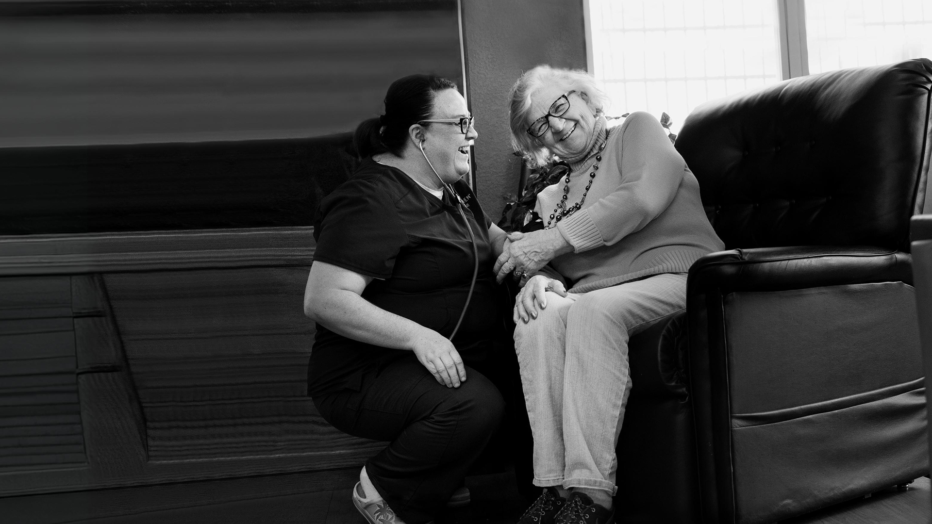 A nurse smiles as she checks the blood pressure of a happy elderly woman sitting on a chair.