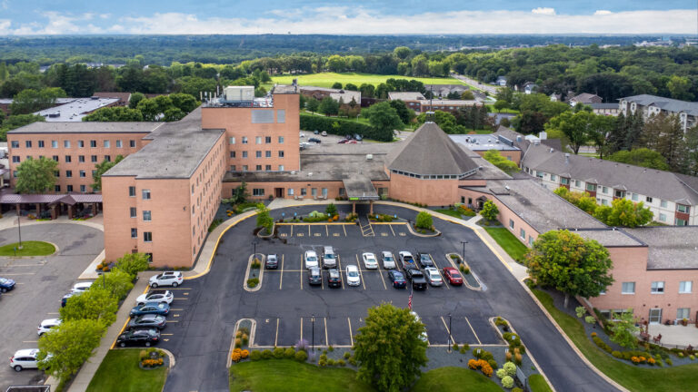 Aerial view of a senior living campus featuring surface parking, interconnected brick buildings and out buildings with nearby park and a partly cloudy sky and horizon in the distance.