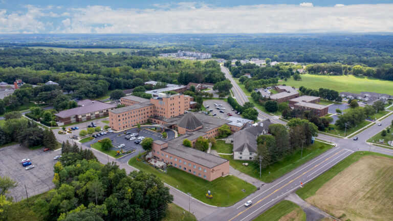 Aerial view of a senior living campus featuring surface parking, interconnected brick buildings and out buildings with nearby primary roads, a park and a partly cloudy sky and horizon in the distance.
