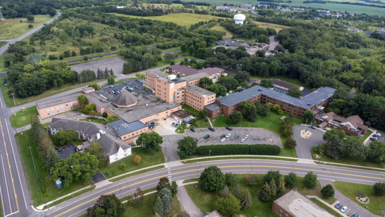 Aerial view of a senior living campus featuring interconnected brick buildings and out buildings with nearby primary roads and a water tower in the distance.