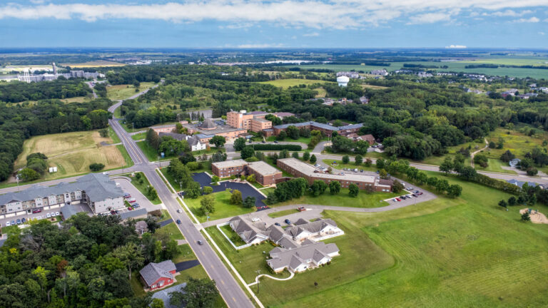 Aerial view of a senior living campus featuring interconnected brick buildings and out buildings with nearby primary roads, water tower and a partly cloudy sky and horizon in the distance.
