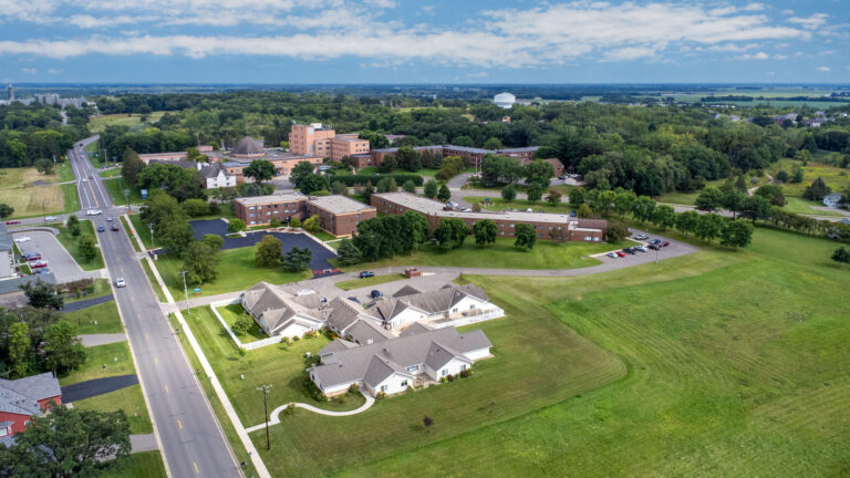 Aerial view of a senior living campus featuring interconnected brick buildings and out buildings with nearby primary roads, water tower and a partly cloudy sky and horizon in the distance.