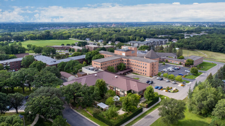 Aerial view of a senior living campus featuring surface parking, interconnected brick buildings, out buildings, and nearby park with partly cloudy sky and horizon in the distance.