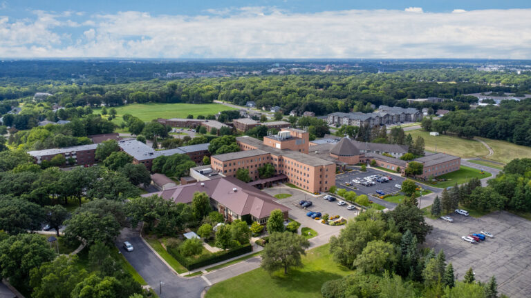 Aerial view of a senior living campus featuring surface parking, interconnected brick buildings, out buildings, and nearby park with partly cloudy sky and horizon in the distance.