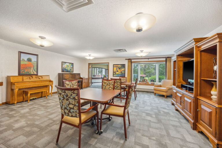 Assisted living community room with tables, chairs, a piano, and a cabinet. Large windows offer views of trees outside.