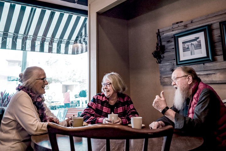 Three older people smiling and talking at a table in a cafe, with coffee cups in front of them.