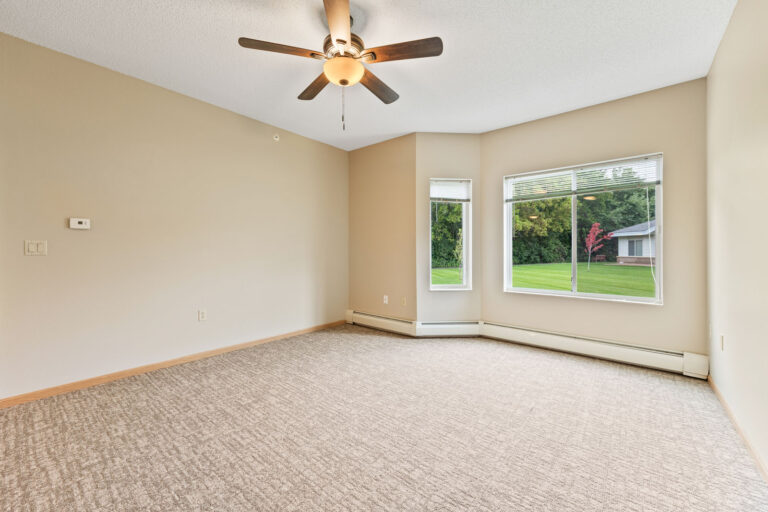Empty beige apartment with carpeted floor, ceiling fan, large windows, and views of lawn and neighboring houses.