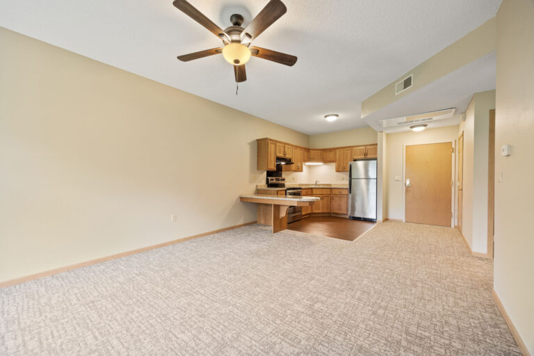 Open living area with ceiling fan, beige carpet, and a kitchen featuring wood cabinets and stainless steel appliances.