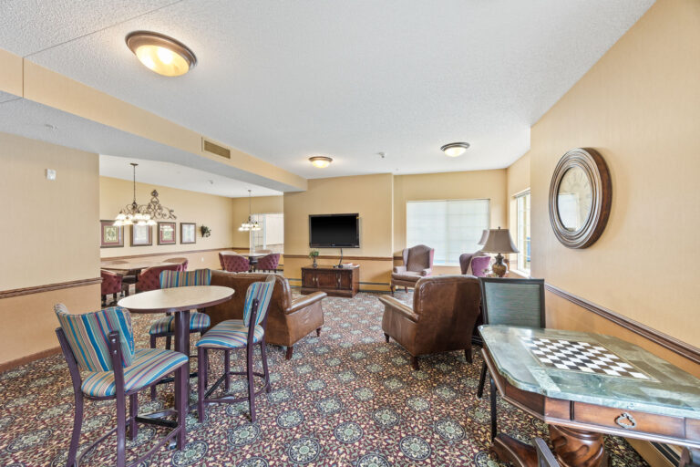 Bright lounge with patterned carpet, chairs, tables, a TV, and large windows letting in natural light.