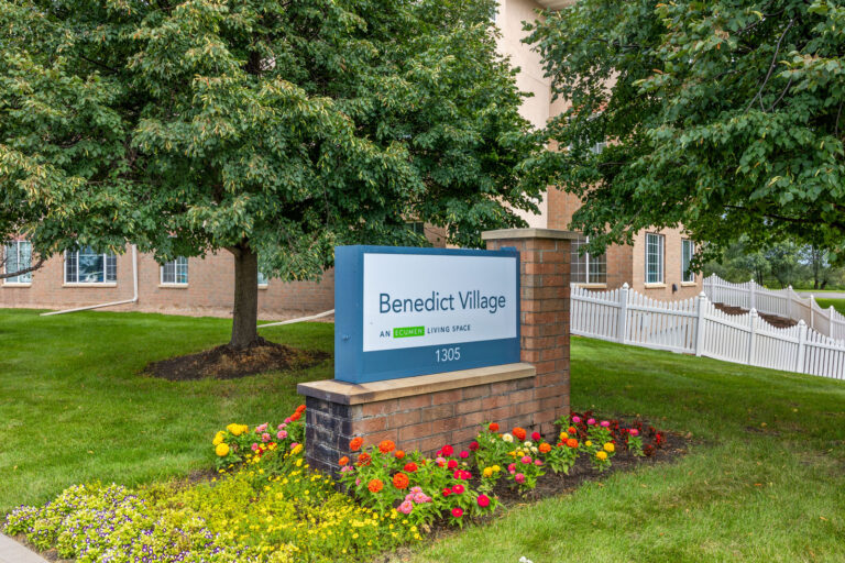 A sign reading Benedict Village stands among flowers and trees in front of a building.