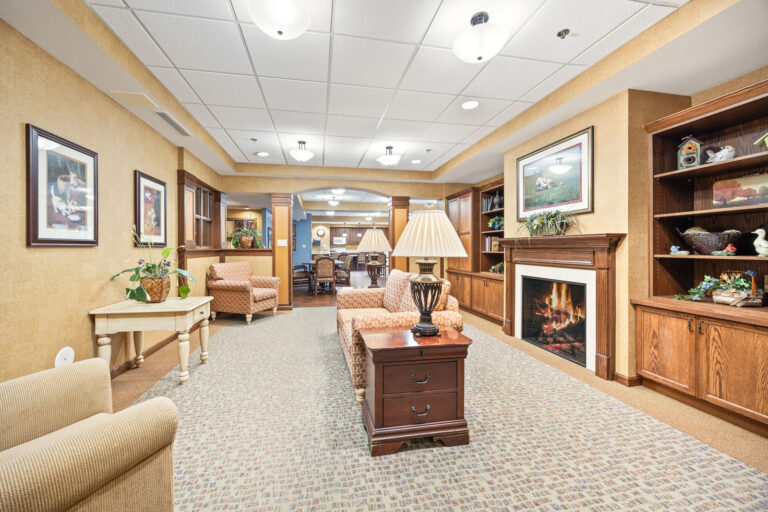 A memory care cozy lounge with armchairs, a sofa, fireplace, bookshelves, art, and warm lighting.